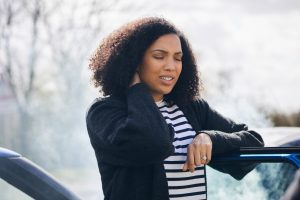 Young woman rubbing neck in pain from whiplash injury standing by damaged car after traffic accident